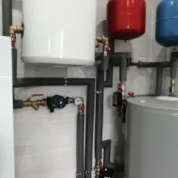 Instalacja centralnego ogrzewania z białym zbiornikiem z logo, czerwonym i niebieskim naczyniem wzbiorczym, szarym zasobnikiem oraz rurami z izolacją termiczną na tle jasnej, wzorzystej ściany.