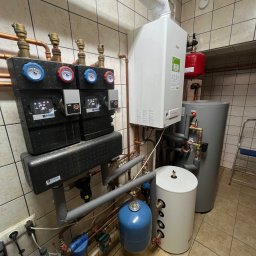 ENERGY INVEST - Kompletna instalacja grzewcza w kotłowni z białym kotłem gazowym, szarym i białym zasobnikiem ciepłej wody, czerwonym naczyniem przeponowym, rozdzielaczami z manometrami oraz niebieskim filtrem wody.