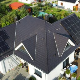 ENERGY INVEST - Widok z góry na dom z ciemną dachówką i panelami fotowoltaicznymi na dachu; ekipa montażowa rozładowuje kolejne panele z samochodu dostawczego.
