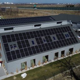 ENERGY INVEST - Widok z góry na szary budynek z panelami słonecznymi na dachu, z oknami dachowymi, z przodu trawnik z niskimi roślinami, obok zaparkowane samochody i śmietniki, w tle pole i autostrada.