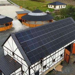 ENERGY INVEST - Widok z góry na budynek stajni z panelami słonecznymi na dachu, obok dwa okrągłe budynki z drewnianymi ścianami i stożkowymi dachami, na ziemi pracują dwie osoby.
