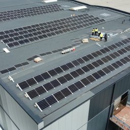 ENERGY INVEST - Montaż paneli słonecznych na dachu hali przemysłowej, widoczni pracownicy w żółtych kamizelkach, częściowo zainstalowane rzędy paneli, palety z panelami czekające na montaż.