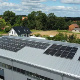 Widok z góry na szary budynek przemysłowy z zamontowanymi panelami słonecznymi na dachu, w tle zabudowania wiejskie i drzewa pod błękitnym, zachmurzonym niebem.