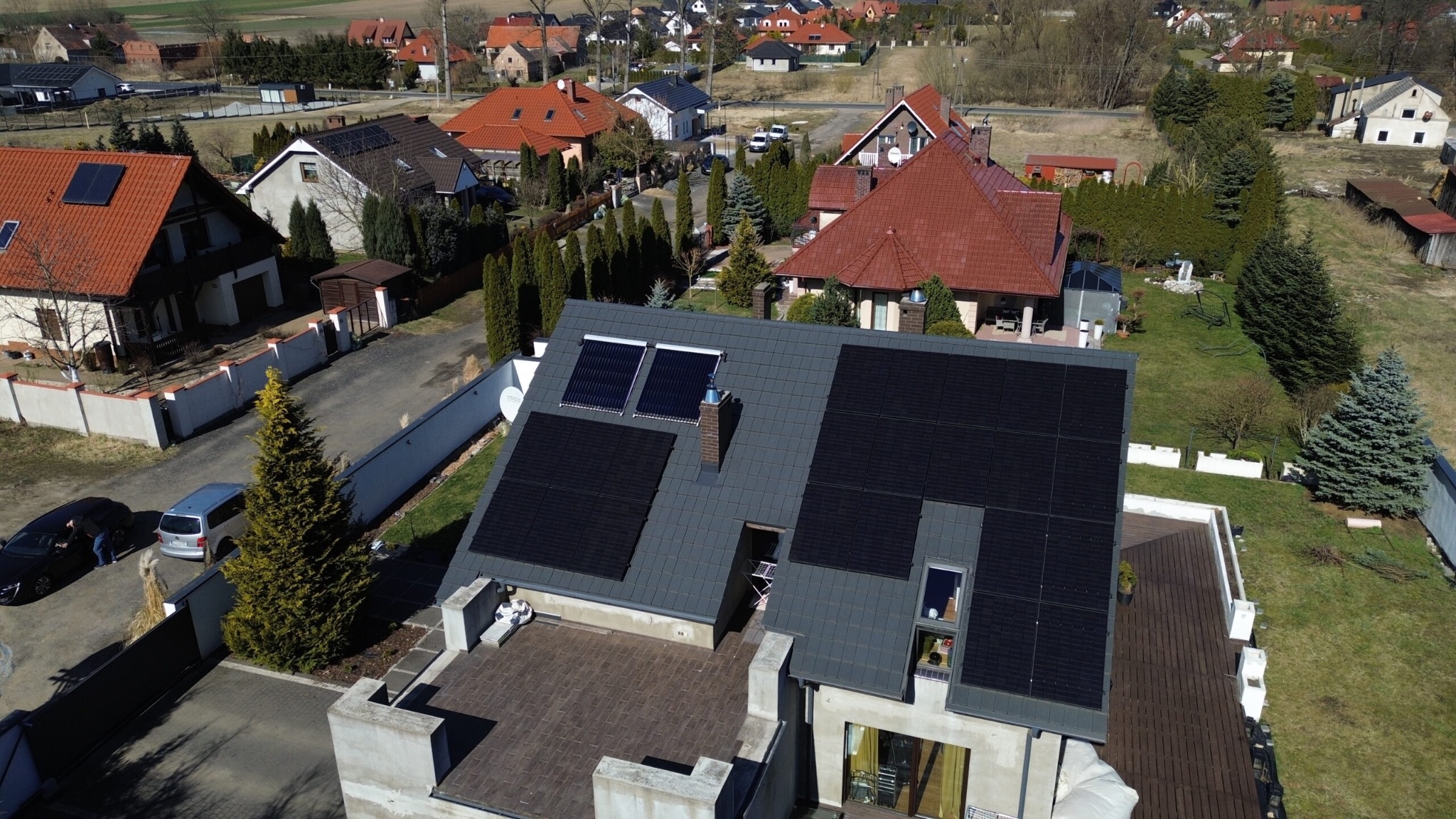 Widok z drona na dom jednorodzinny z szarym dachem, na którym zamontowano czarne panele fotowoltaiczne, oraz dodatkowo dwa kolektory słoneczne; w tle zabudowa wiejska.
