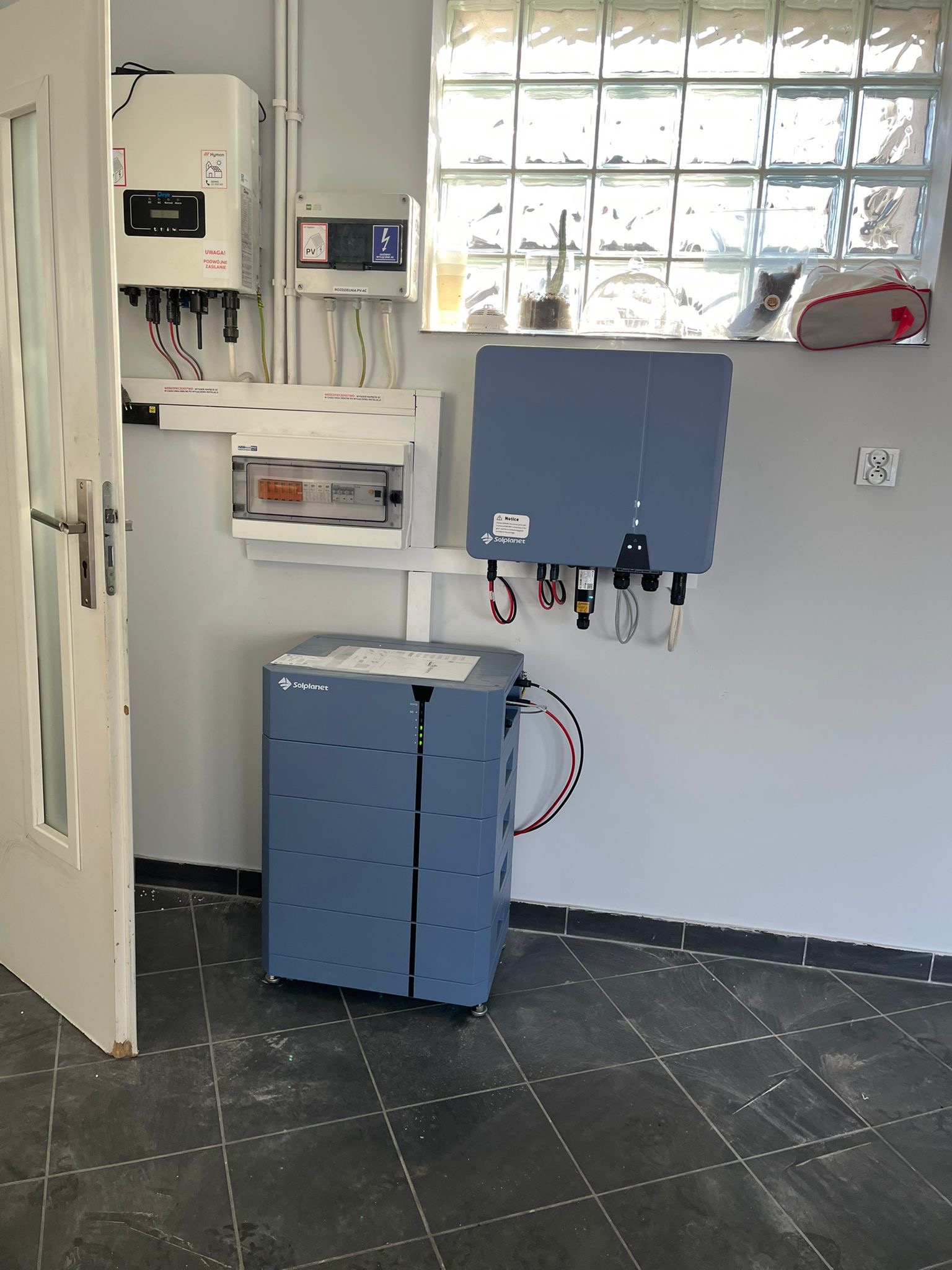 Kompletny zestaw instalacji domowej: falownik, rozdzielnica elektryczna, moduł sterujący i bateria magazynu energii Solplanet, umieszczone w pomieszczeniu z oknem z luksferów i ciemną, kwadratową...