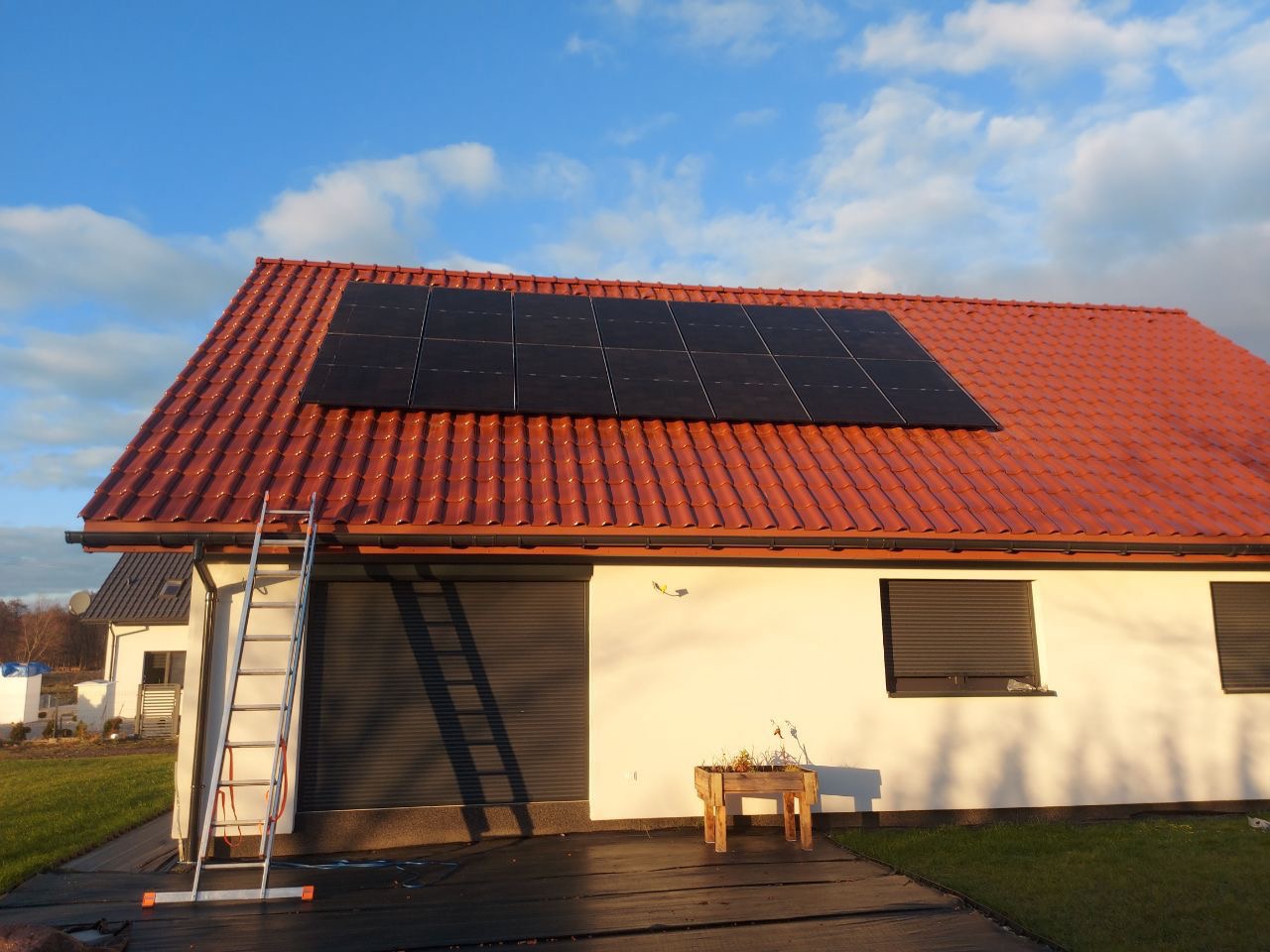 Czerwony dach z panelami słonecznymi w układzie prostokątnym, aluminiowa drabina oparta o ścianę budynku, błękitne niebo z chmurami w tle.