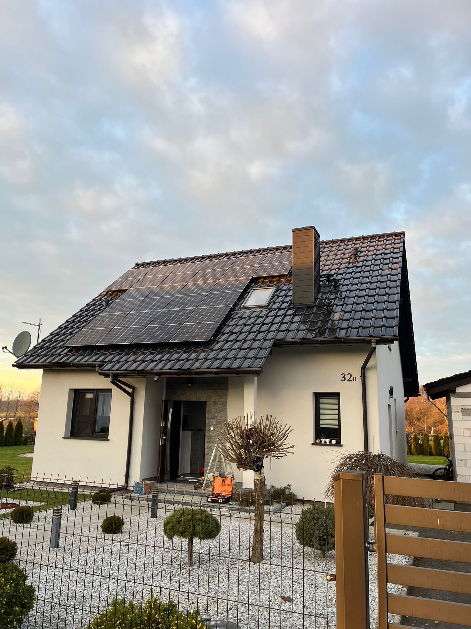 Biały dom jednorodzinny z ciemną dachówką i panelami słonecznymi na dachu, widoczne drabiny i skrzynka narzędziowa sugerują trwający montaż, ogród z ozdobnymi krzewami i ogrodzeniem.