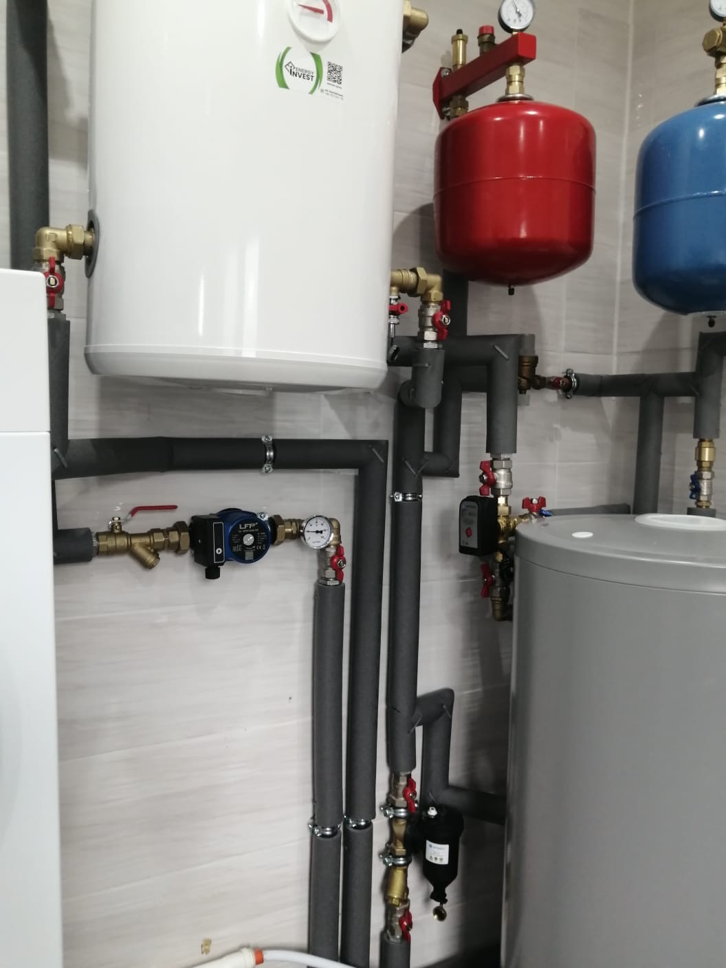 Instalacja centralnego ogrzewania z białym zbiornikiem z logo, czerwonym i niebieskim naczyniem wzbiorczym, szarym zasobnikiem oraz rurami z izolacją termiczną na tle jasnej, wzorzystej ściany.