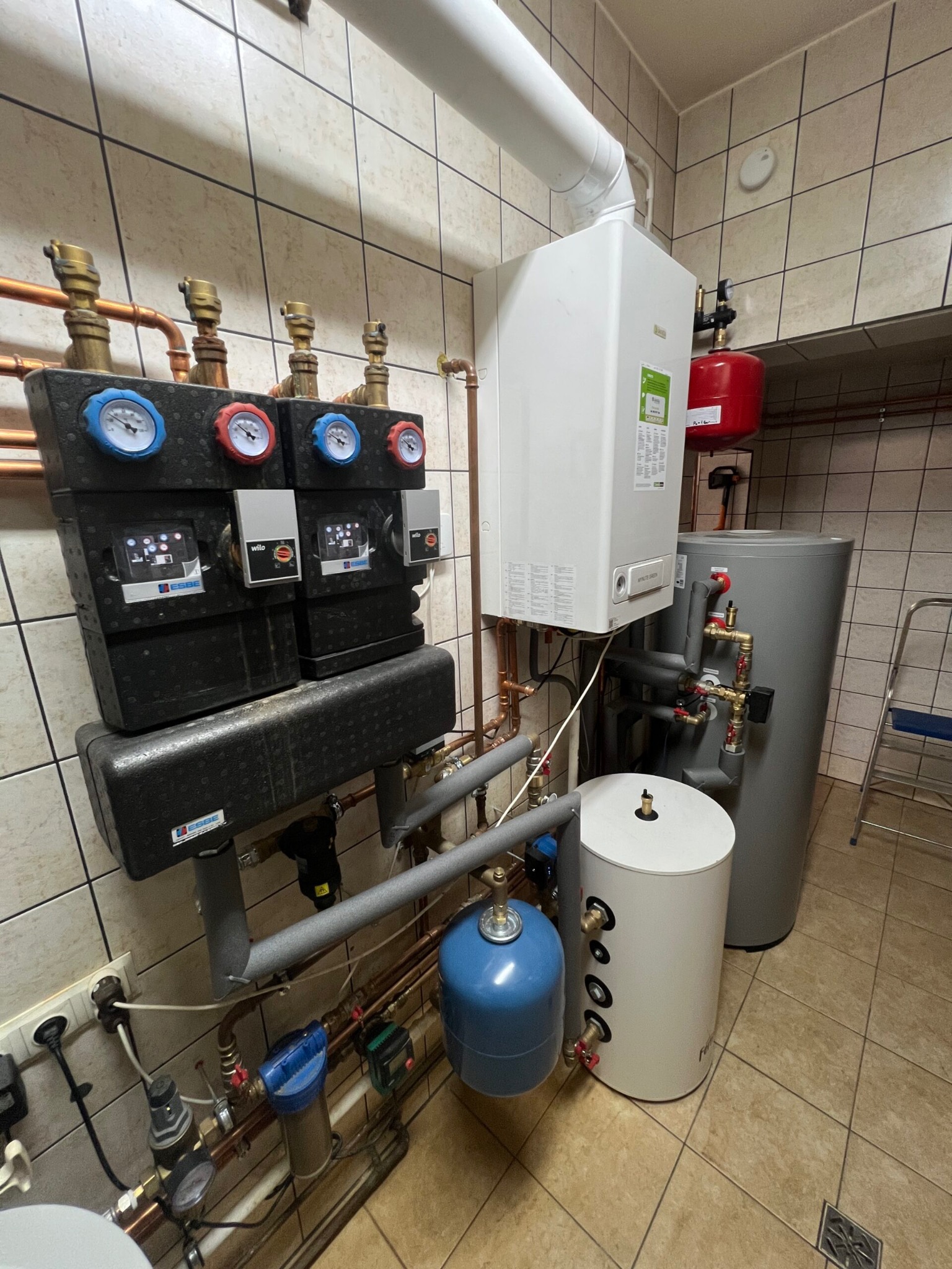 Kompletna instalacja grzewcza w kotłowni z białym kotłem gazowym, szarym i białym zasobnikiem ciepłej wody, czerwonym naczyniem przeponowym, rozdzielaczami z manometrami oraz niebieskim filtrem wody.
