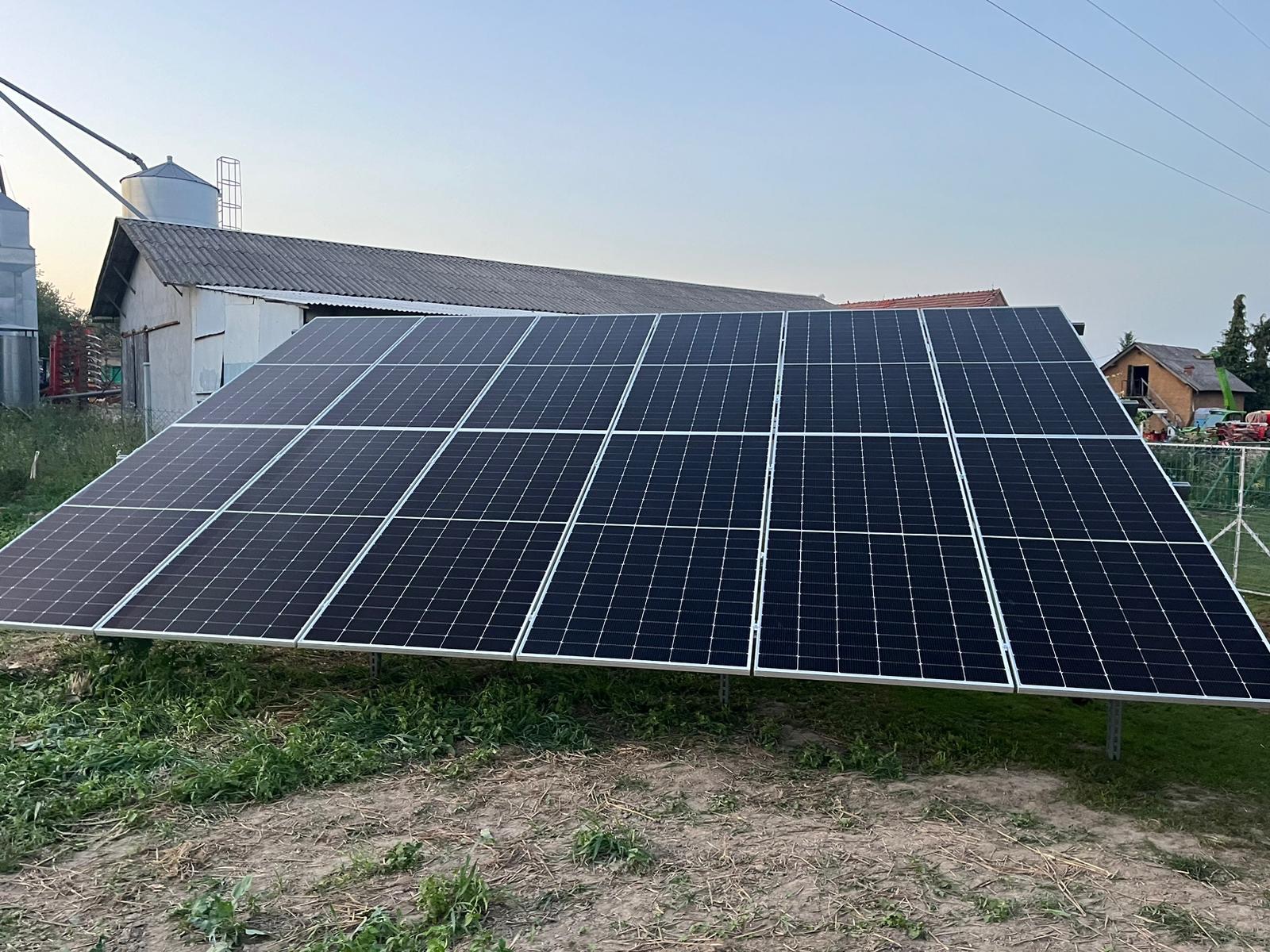 Panel fotowoltaiczny na metalowej konstrukcji, z budynkami gospodarczymi i silosem w tle, w wiejskim krajobrazie.
