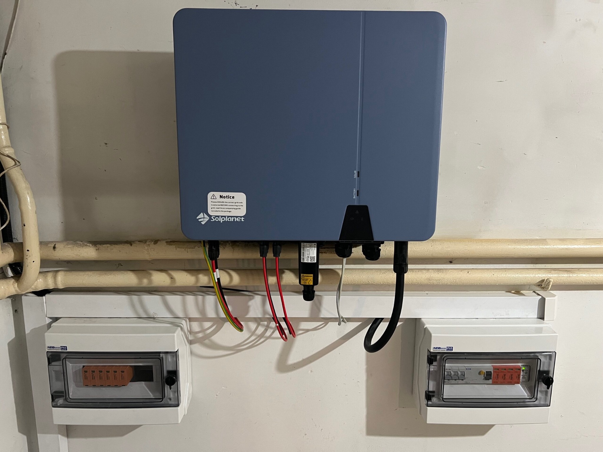 Niebieski inwerter z naklejką ostrzegawczą Solplanet, podłączony przewodami do dwóch białych skrzynek z bezpiecznikami na ścianie. Widoczne rury instalacyjne w tle.