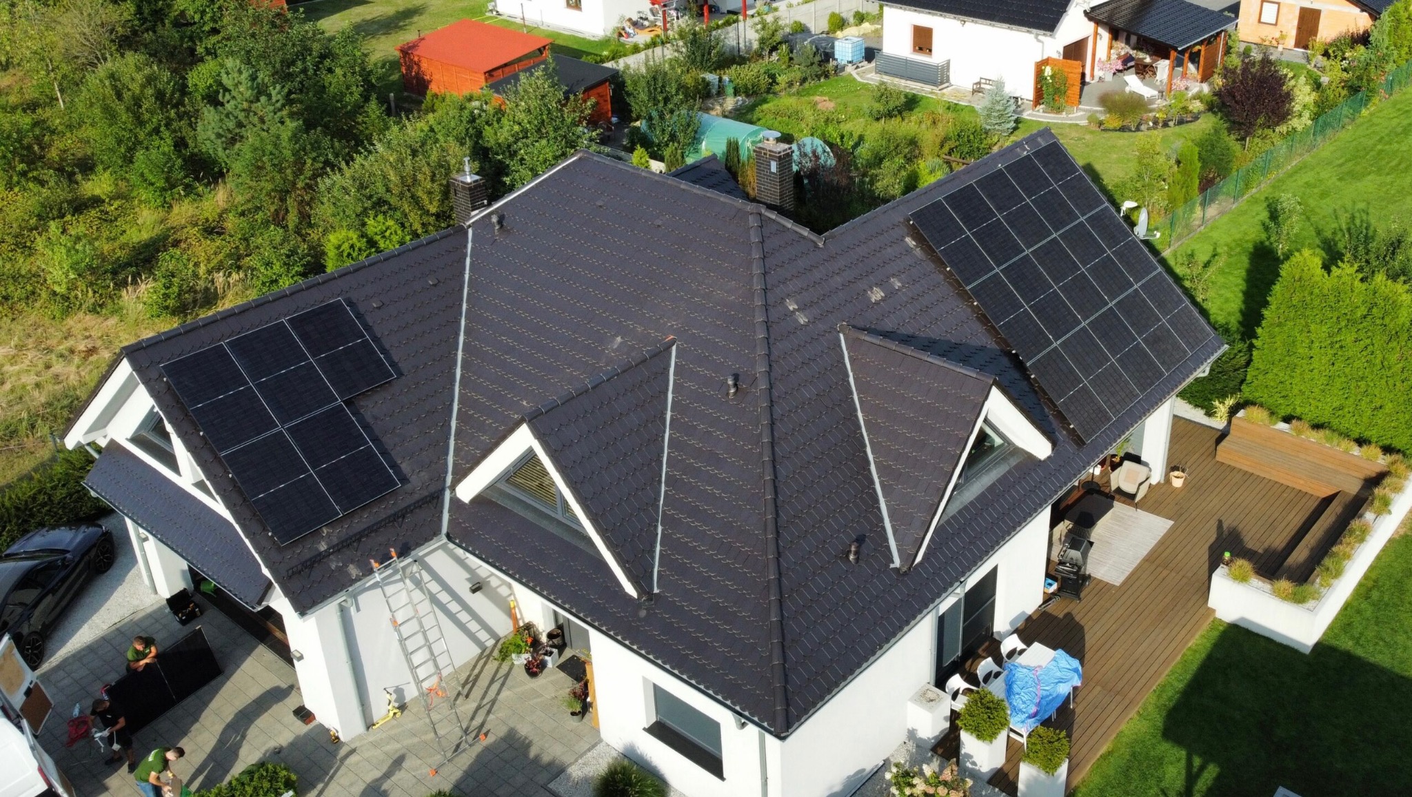 Widok z góry na dom z ciemną dachówką i panelami fotowoltaicznymi na dachu; ekipa montażowa rozładowuje kolejne panele z samochodu dostawczego.