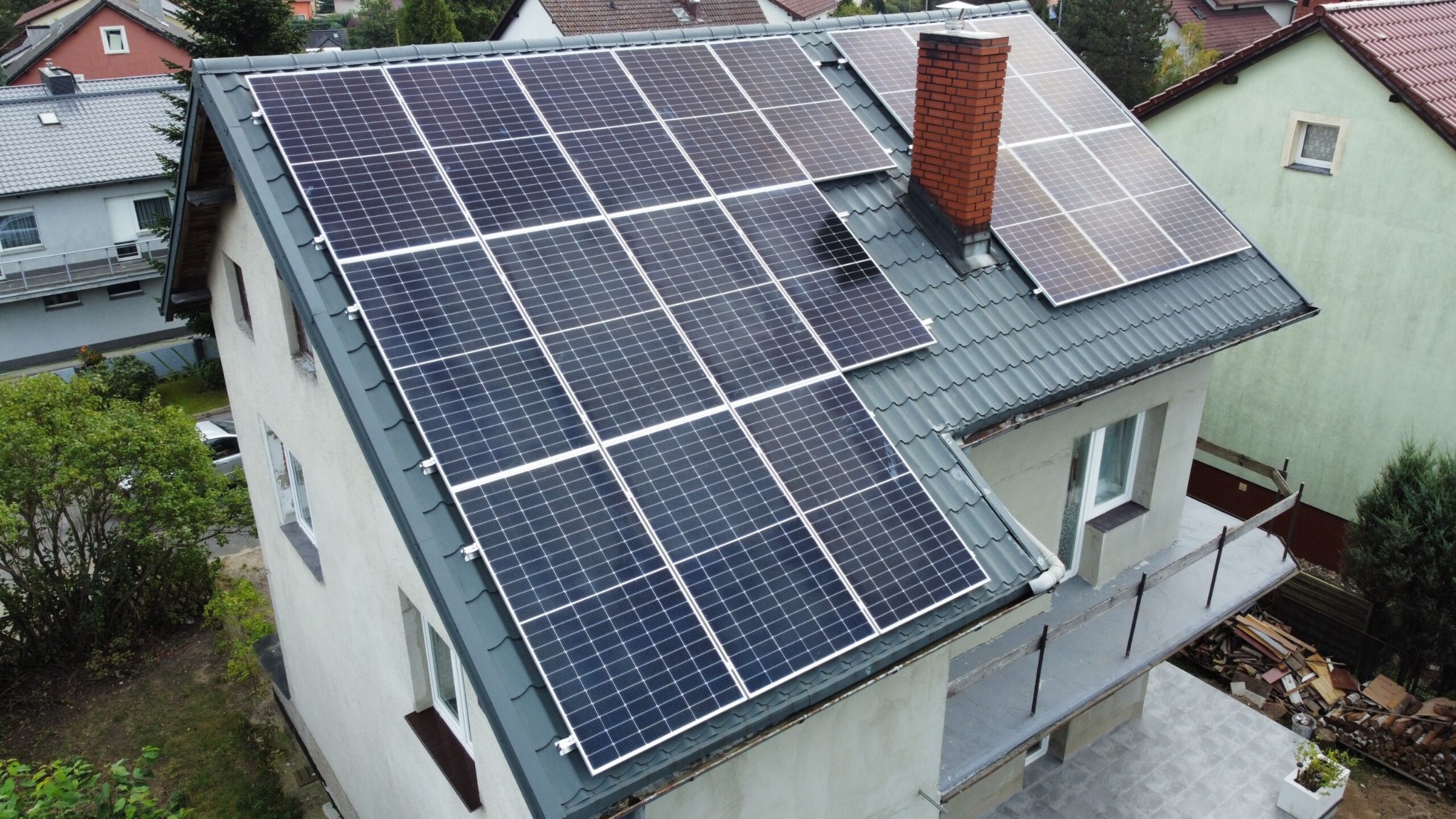 Widok z góry na dom jednorodzinny z szarym dachem, na którym zamontowane są panele słoneczne; obok komina umieszczone są dodatkowe panele.