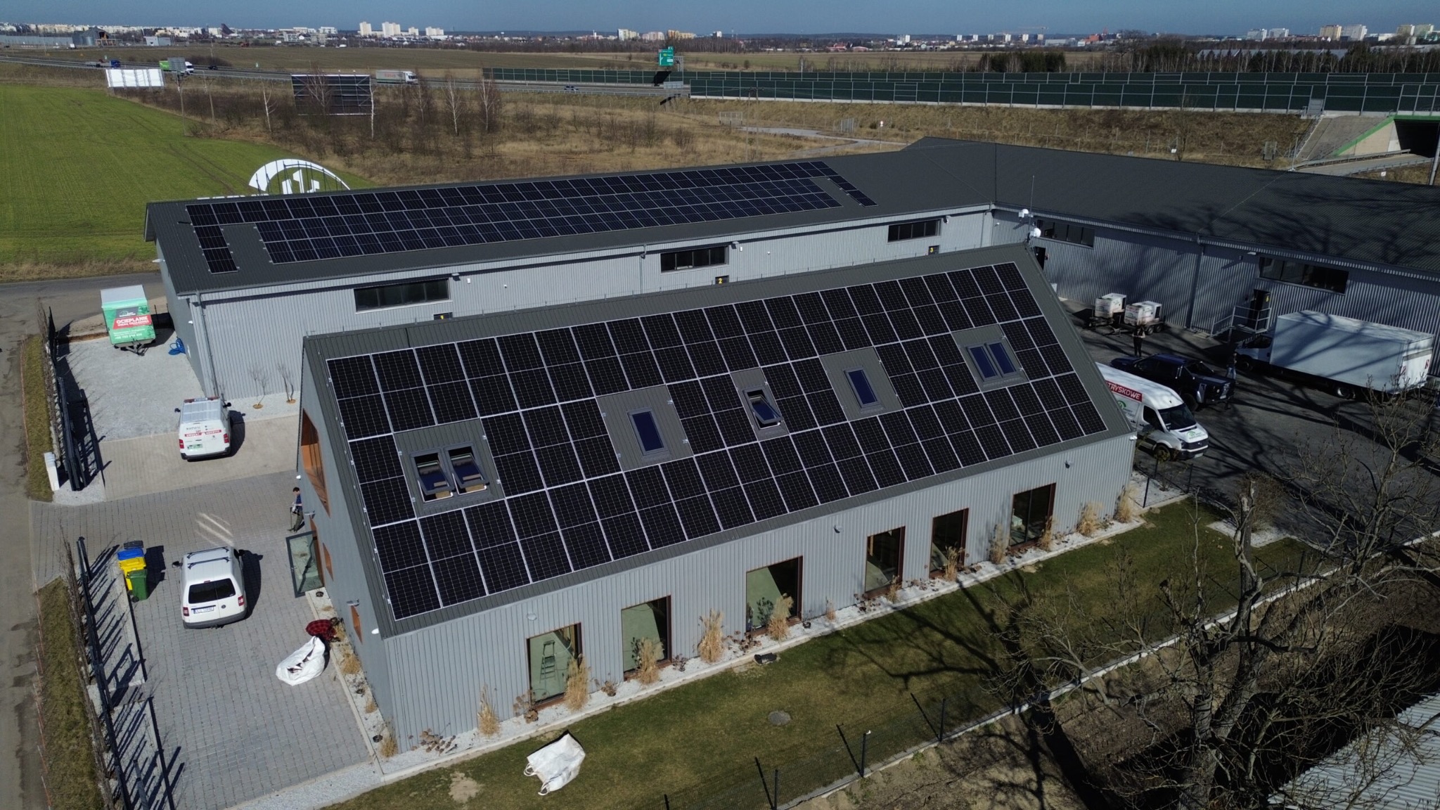 Widok z góry na szary budynek z panelami słonecznymi na dachu, z oknami dachowymi, z przodu trawnik z niskimi roślinami, obok zaparkowane samochody i śmietniki, w tle pole i autostrada.