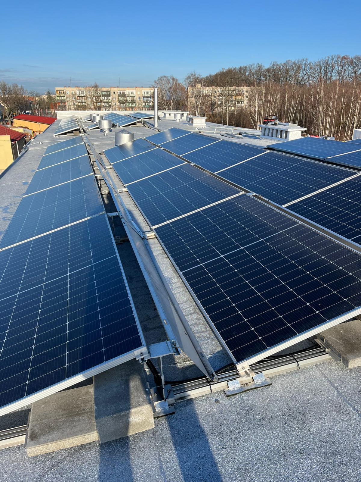 Rząd paneli słonecznych na płaskim dachu budynku z widocznymi elementami montażowymi i budynkami w tle pod błękitnym niebem.