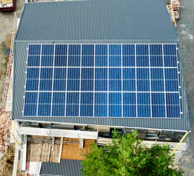 Widok z góry na budynek z szarym dachem krytym blachą falistą, na którym zamontowano panel słoneczny składający się z wielu ogniw, otoczony rusztowaniami i gruzem, z zielenią drzew w dolnej części...