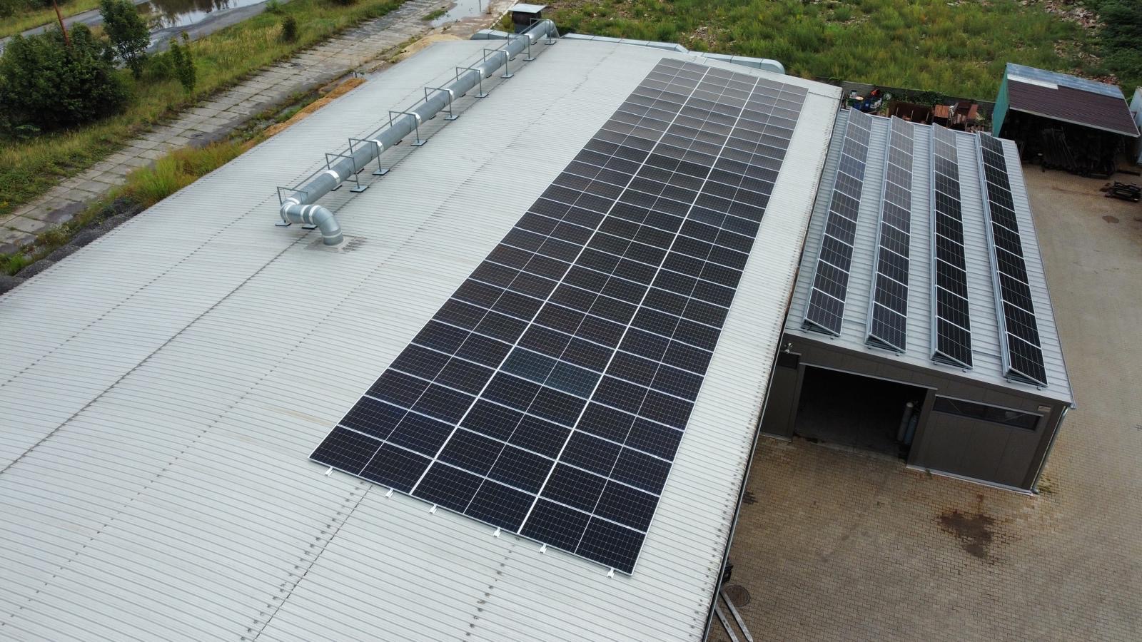 Widok z góry na instalację paneli słonecznych na dachu budynku przemysłowego z widocznymi elementami wentylacji i sąsiednim budynkiem z dodatkowymi panelami.