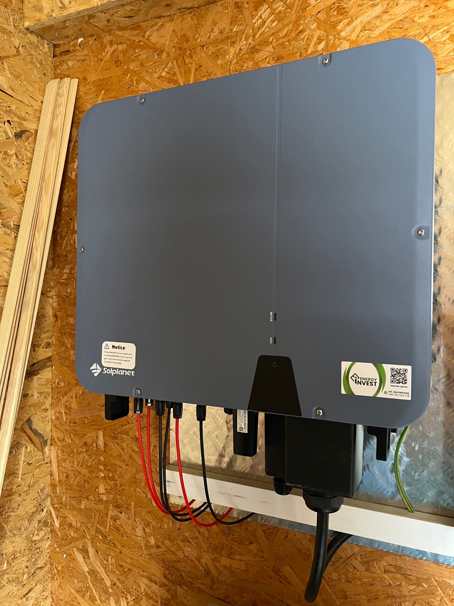 Szary falownik Solplanet zamontowany na ścianie z płyty OSB, z widocznymi kablami i naklejką z logo Energy Invest oraz kodem QR do wystawienia opinii.