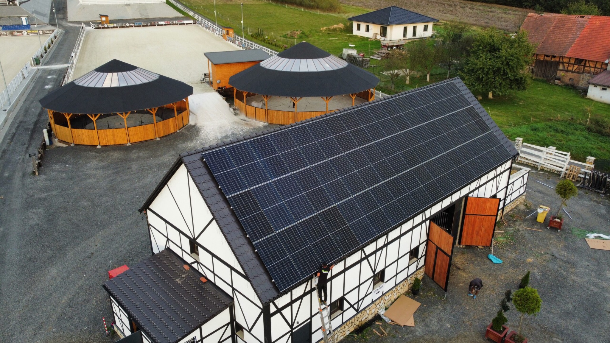 Widok z góry na budynek stajni z panelami słonecznymi na dachu, obok dwa okrągłe budynki z drewnianymi ścianami i stożkowymi dachami, na ziemi pracują dwie osoby.