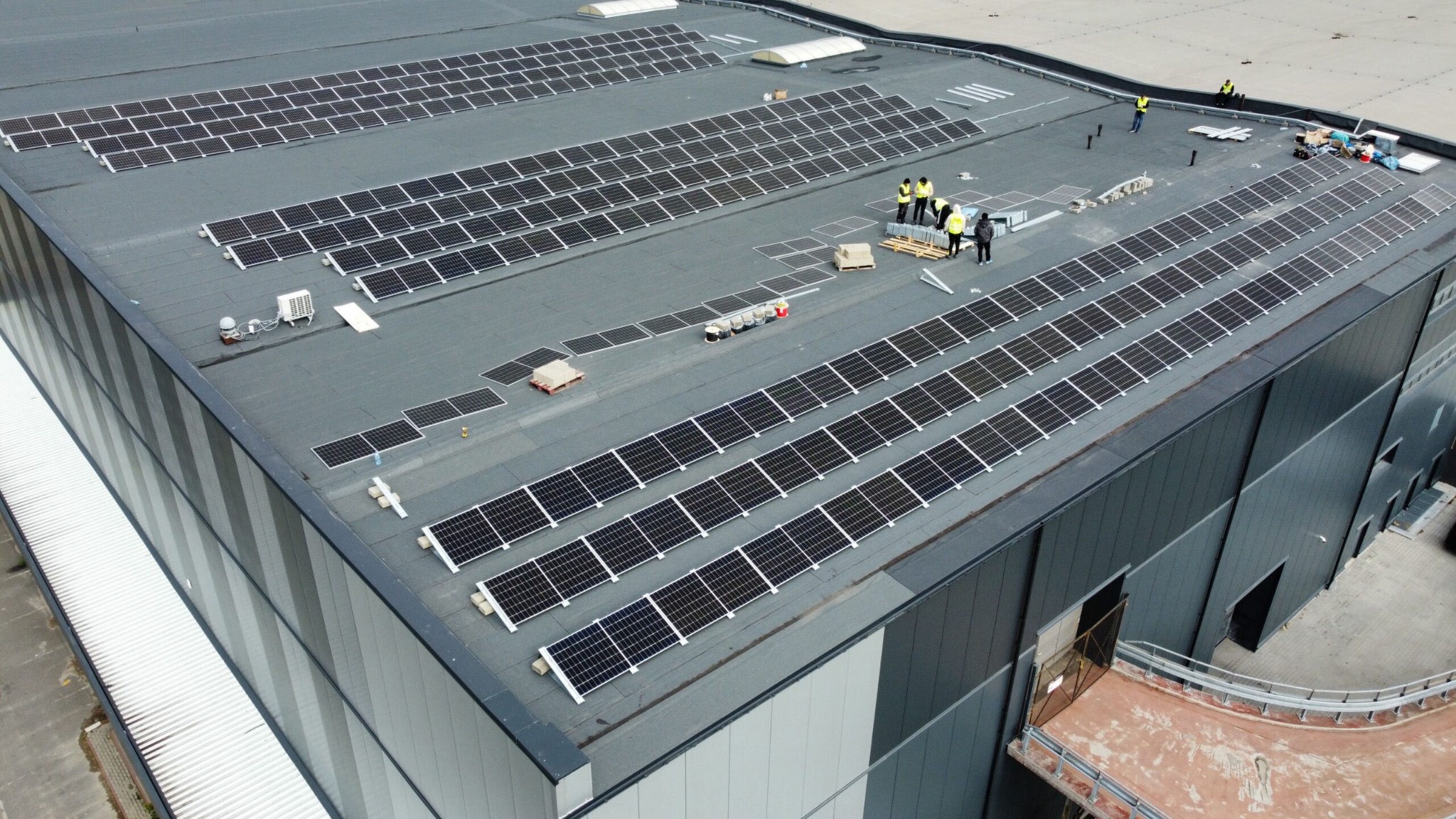 Montaż paneli słonecznych na dachu hali przemysłowej, widoczni pracownicy w żółtych kamizelkach, częściowo zainstalowane rzędy paneli, palety z panelami czekające na montaż.