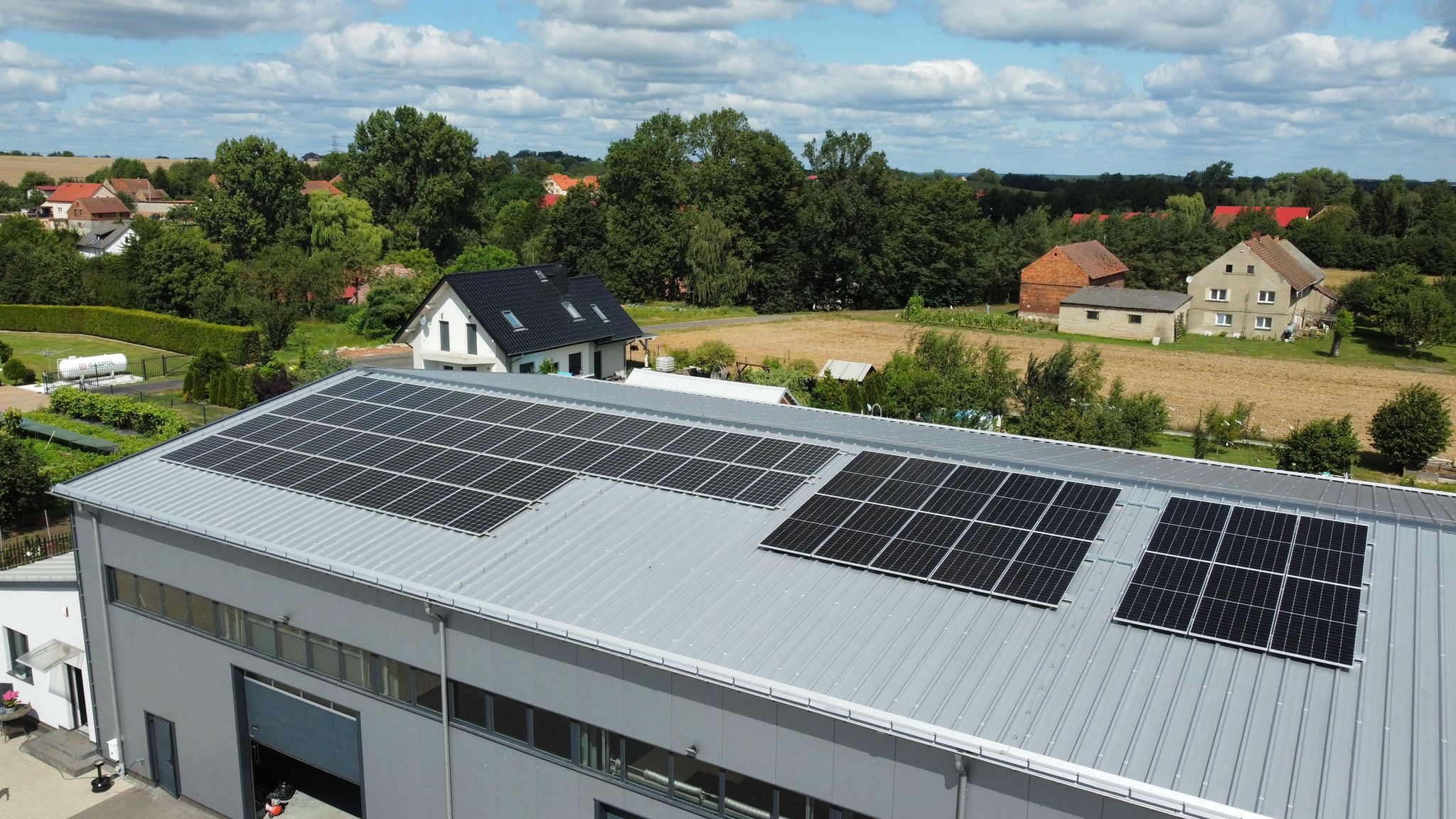 Widok z góry na szary budynek przemysłowy z zamontowanymi panelami słonecznymi na dachu, w tle zabudowania wiejskie i drzewa pod błękitnym, zachmurzonym niebem.