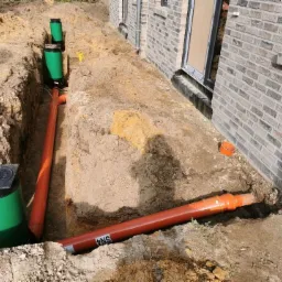 Wykop z ułożonymi rurami kanalizacyjnymi i studzienkami rewizyjnymi przy ścianie budynku z cegły. Widoczne pomarańczowe rury PCV i zielone studzienki z czarnymi pokrywami. Na jednej z rur widoczny...