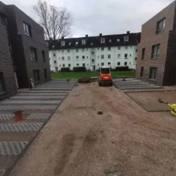 Teren pomiędzy nowymi budynkami z kostką ażurową w trakcie układania, widoczny mały walec i minikoparka.