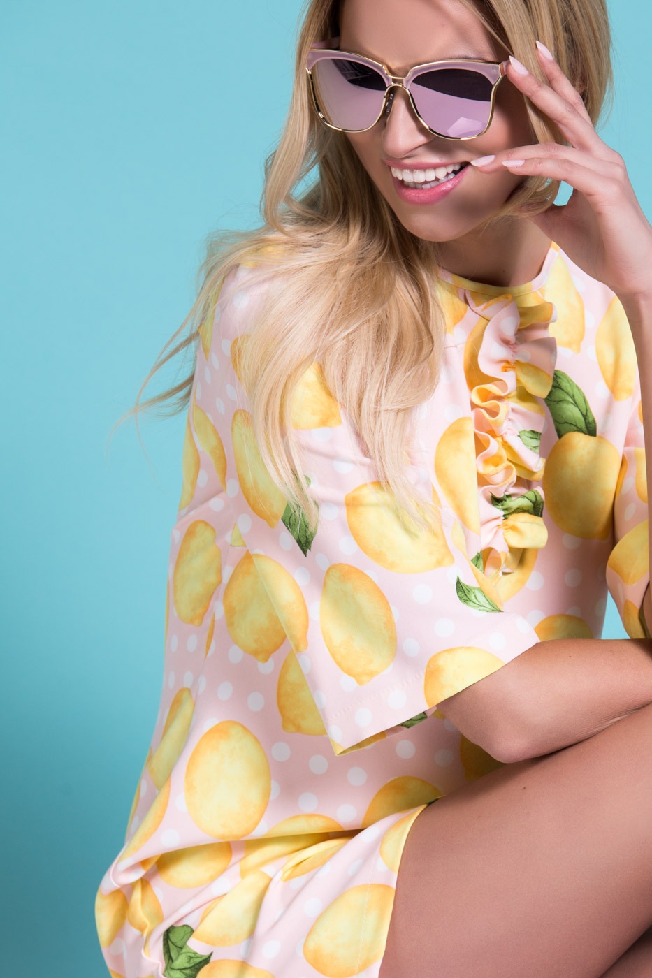 Uśmiechnięta blondynka w okularach przeciwsłonecznych, ubrana w różową bluzkę w cytryny i białe kropki, dotyka palcami policzka na tle turkusowego studia.