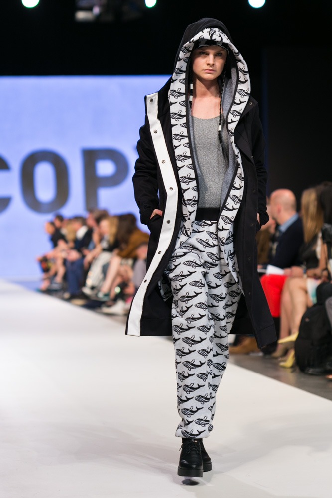 Modelka prezentuje czarny płaszcz z białym wykończeniem i spodnie w czarno-białe wzory z motywem ryb na wybiegu podczas pokazu mody, widoczna publiczność w tle.