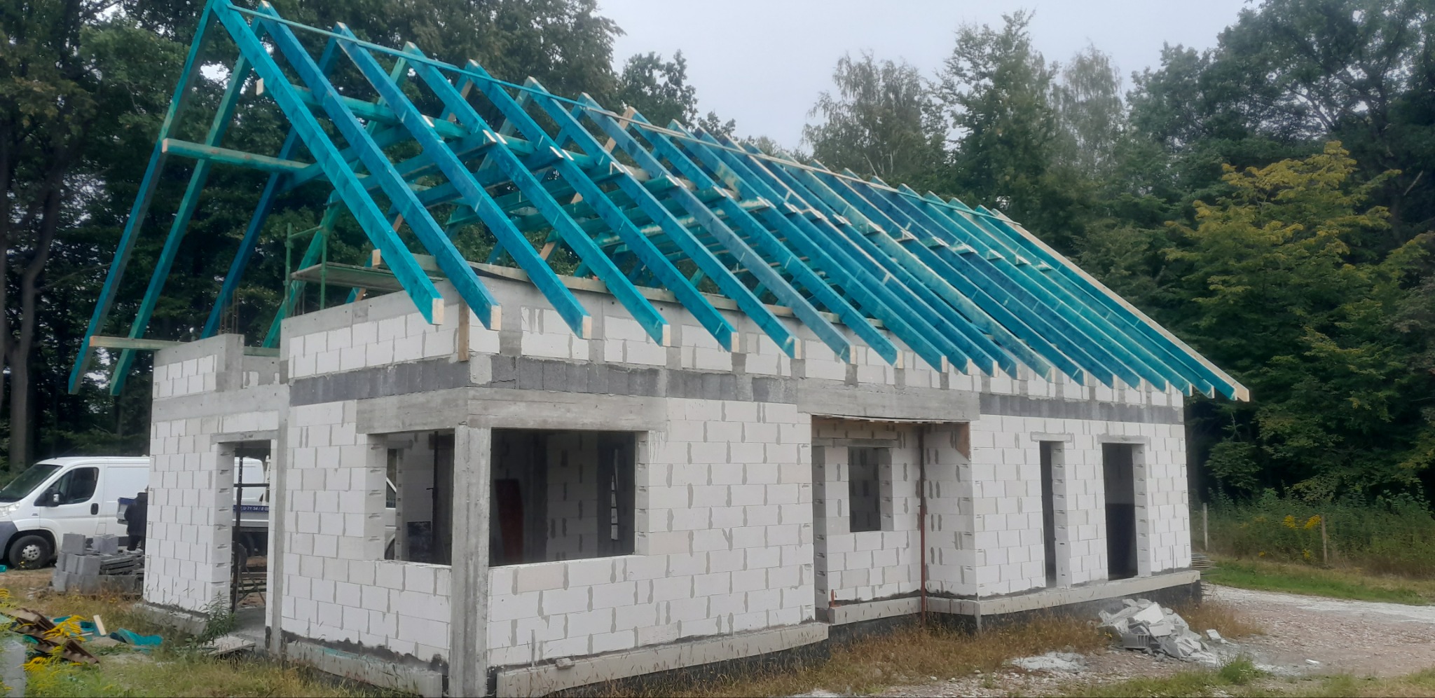Surowy stan domu murowanego z niebieską konstrukcją dachu w lesistej okolicy, widoczny biały van.
