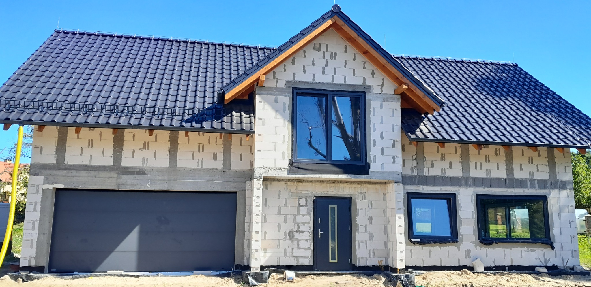Surowy stan domu z białego bloczka, z zamontowanymi oknami w czarnych ramach i grafitową dachówką, widoczny z zewnątrz w słoneczny dzień.