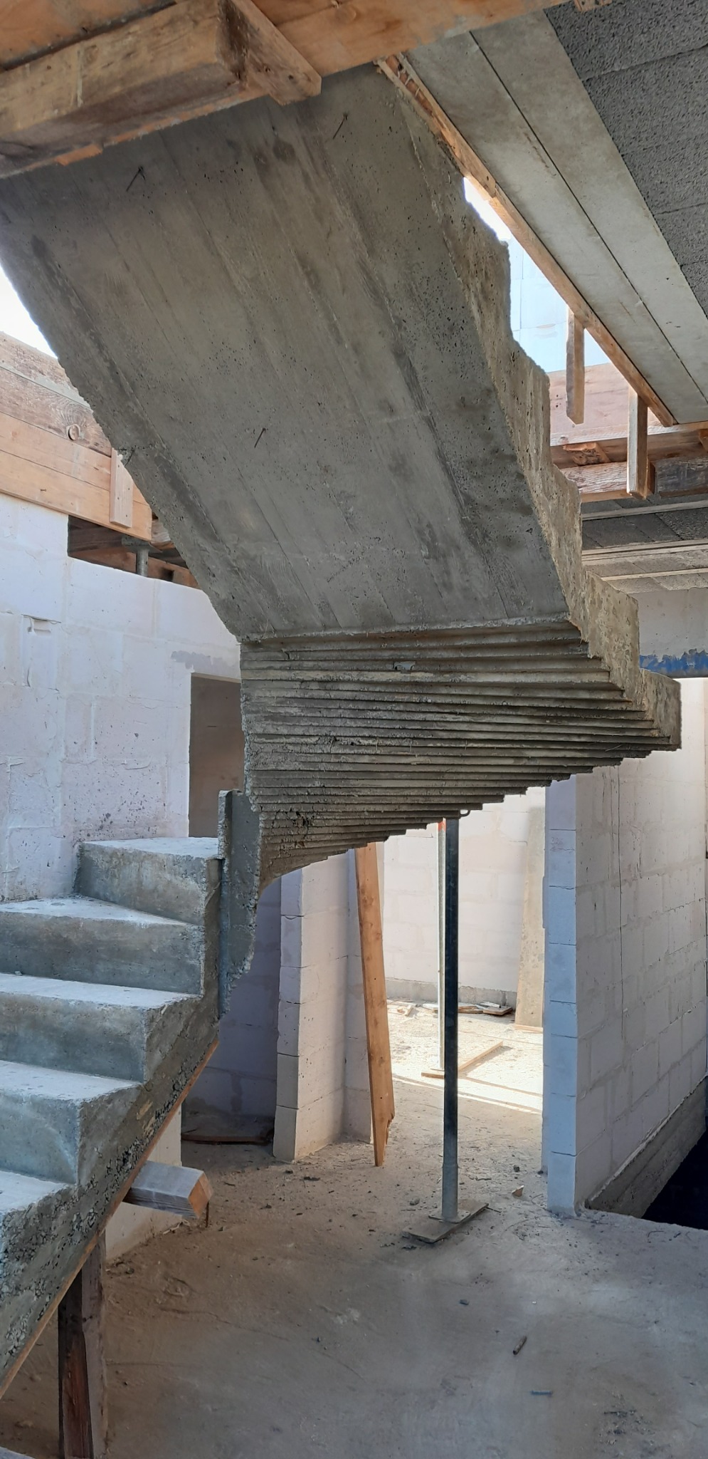 Surowe betonowe schody wewnętrzne w trakcie budowy, widoczne drewniane szalunki i podpory, ściany z białych bloczków.