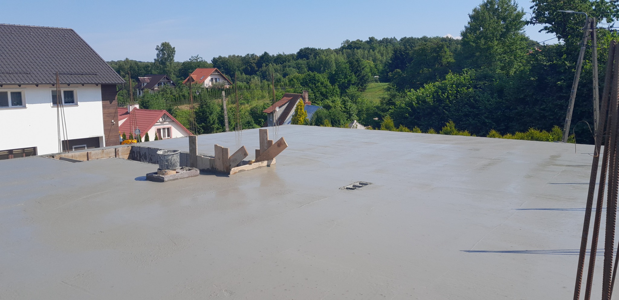 Świeżo wylany, gładki betonowy strop na budowie domu jednorodzinnego, widoczne elementy szalunków i wystające pręty zbrojeniowe, w tle zabudowa mieszkalna i zieleń.
