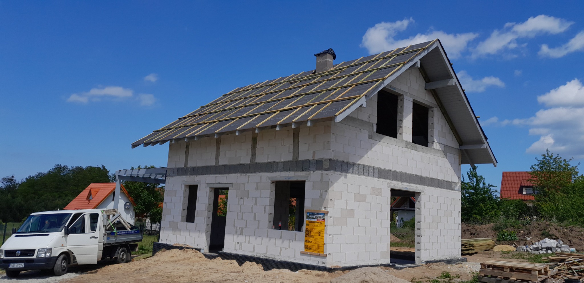 Niedokończony dom z białego pustaka, w trakcie budowy dachu z widoczną konstrukcją drewnianą i czarną membraną, na tle błękitnego nieba z chmurami, obok zaparkowany biały samochód dostawczy.