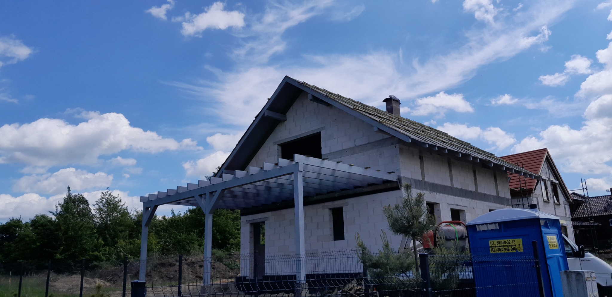 Niedokończony dom murowany z szarej cegły z drewnianą pergolą, widoczny fragment dachu z deskami i kominem oraz przenośna toaleta na placu budowy.