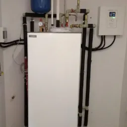 Biała centrala wentylacyjna z niebieskim zbiornikiem wyrównawczym, rurami i panelem sterowania zamontowana na ścianie w pomieszczeniu technicznym.