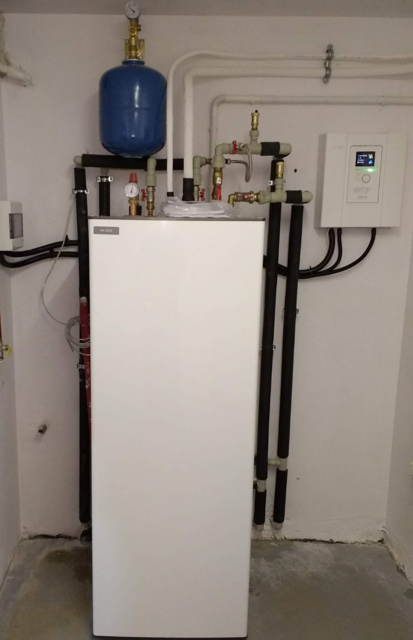 Biała centrala wentylacyjna z niebieskim zbiornikiem wyrównawczym, rurami i panelem sterowania zamontowana na ścianie w pomieszczeniu technicznym.