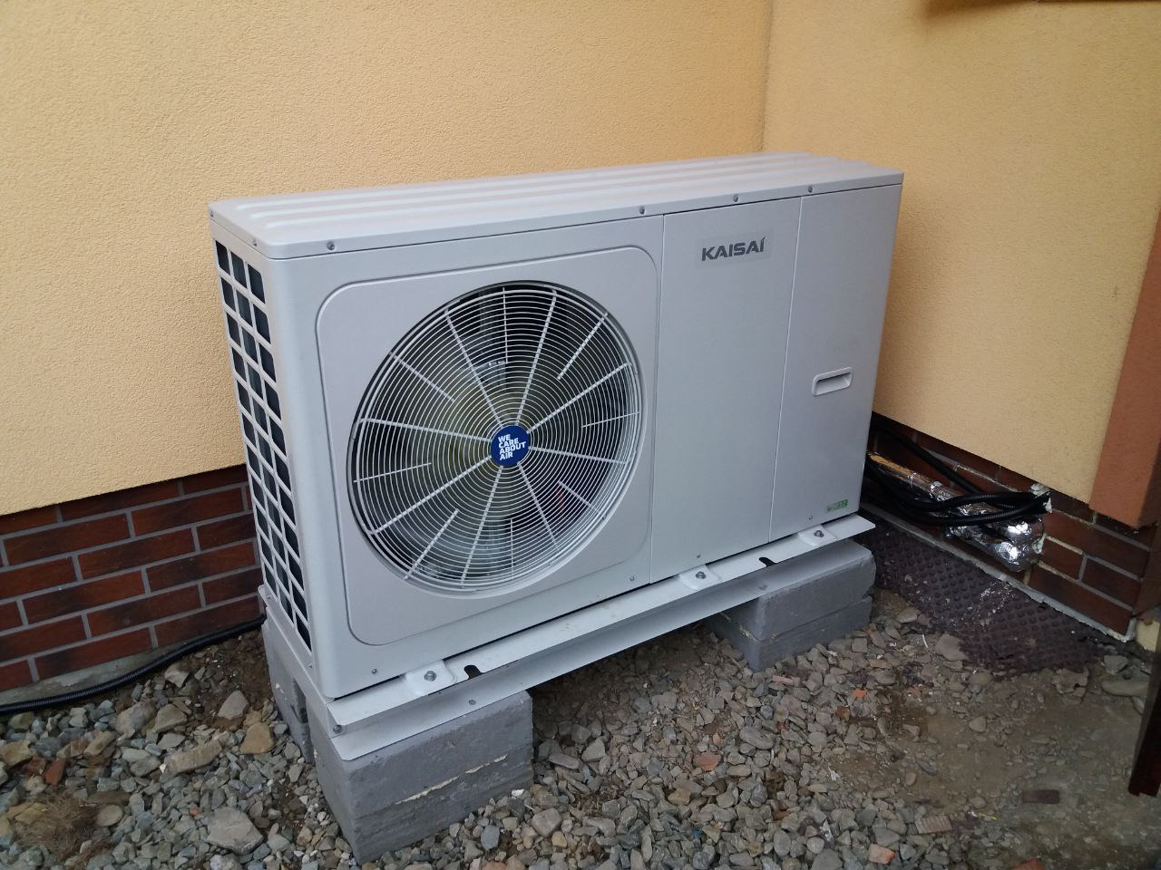 Szara zewnętrzna jednostka klimatyzacji marki Kaisai, zamontowana na betonowych bloczkach przy ścianie budynku o żółtej elewacji i ceglanej podmurówce, widoczne przewody w izolacji.