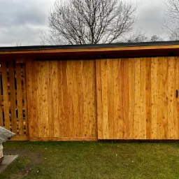 Drewniany schowek ogrodowy z przesuwnymi drzwiami, widoczny na tle trawnika i nagiego drzewa w pochmurny dzień.