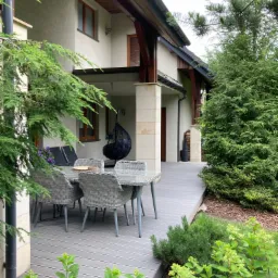 Szary taras z desek kompozytowych przy domu, z szarymi meblami ogrodowymi i wiszącym fotelem kokonem, otoczony zielenią.