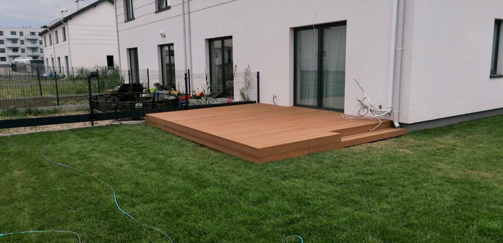 Nowoczesny, brązowy taras z desek kompozytowych z dwoma stopniami, przylegający do białego budynku z dużymi oknami, widoczny trawnik i ogrodzenie.