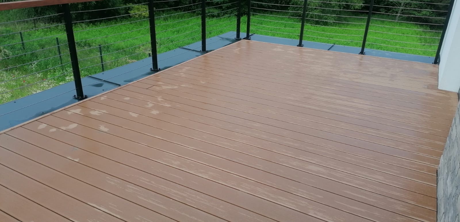 Brązowy taras z desek kompozytowych, z czarną balustradą z linkami stalowymi, widok na zielony trawnik i drzewa.