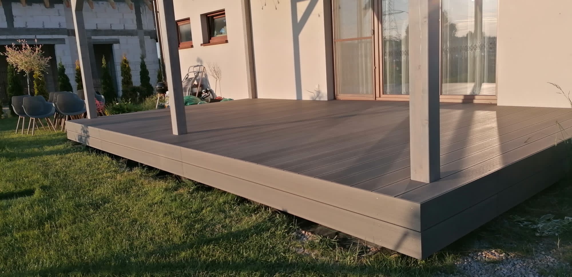 Nowoczesny taras kompozytowy w kolorze szarości, z widocznymi słupami konstrukcyjnymi, przylegający do domu z oknami i drzwiami tarasowymi, otoczony zielonym trawnikiem.
