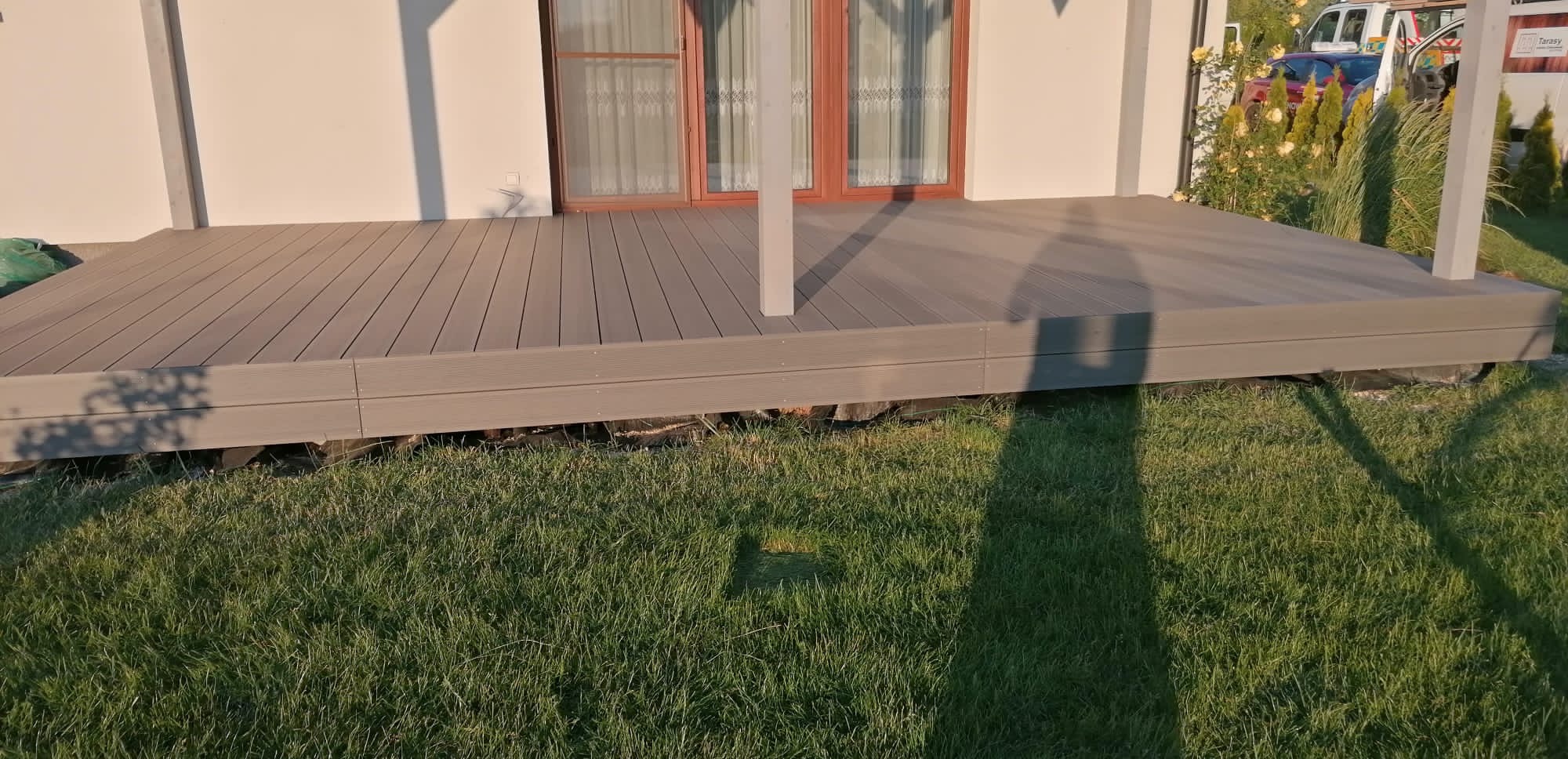 Szary taras z desek kompozytowych przy białym domu z brązowymi oknami, widok z poziomu trawnika, cień osoby na trawie.