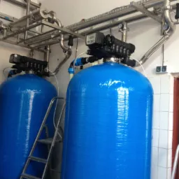 Dwa niebieskie zbiorniki ciśnieniowe z czarnymi głowicami sterującymi, połączone rurami ze stali nierdzewnej, ustawione w pomieszczeniu z białą glazurą i drabiną inspekcyjną.