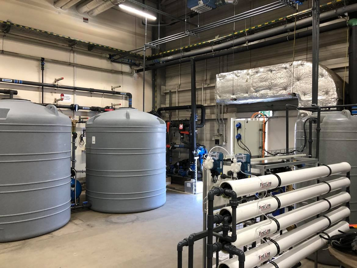 Wnętrze przemysłowej instalacji z dwoma dużymi, szarymi zbiornikami, systemem rur i urządzeń filtrujących z białymi cylindrami oznaczonymi 'First Line', oświetleniem sufitowym i metalową izolacją...