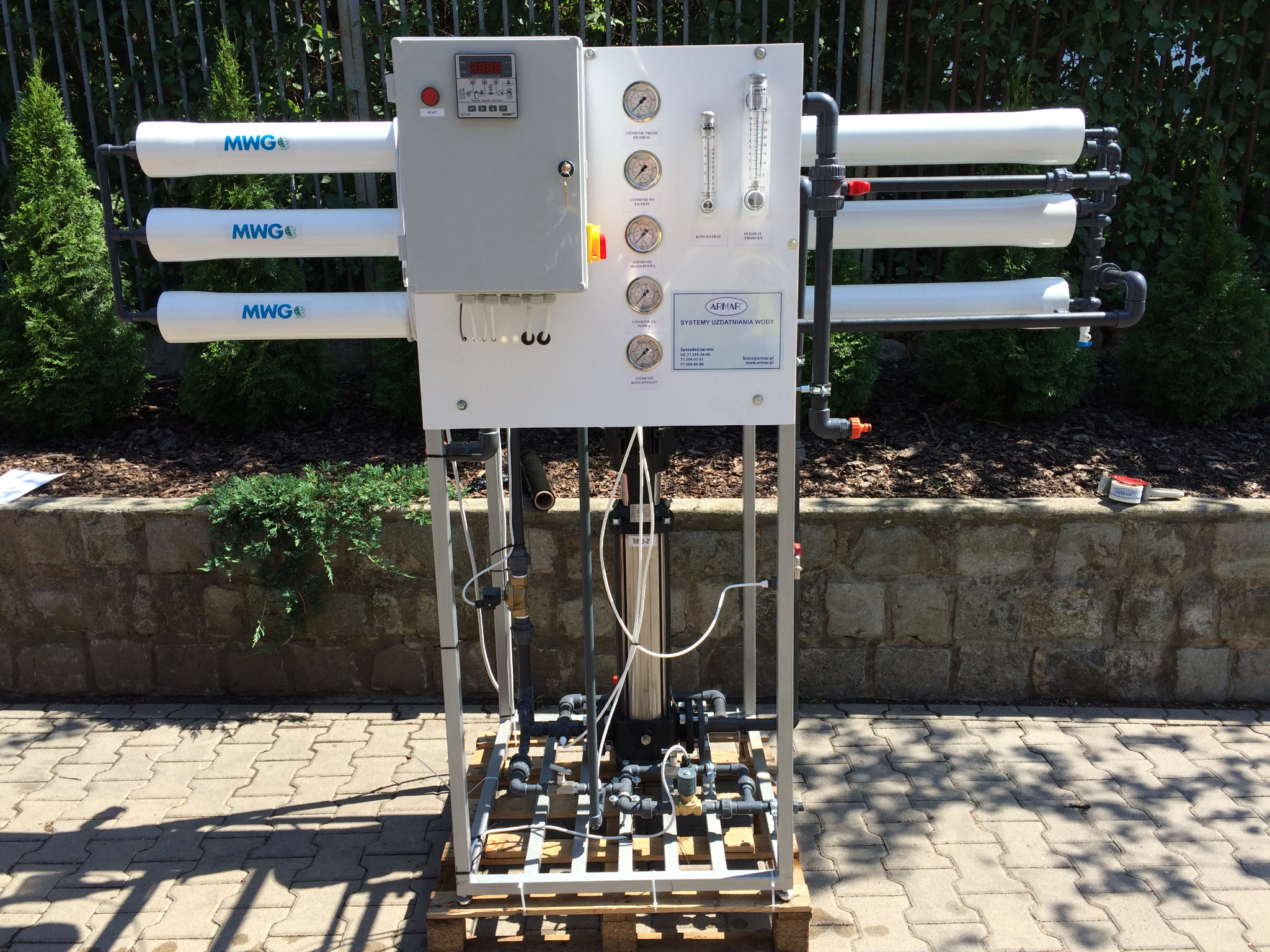 Kompletny system uzdatniania wody z trzema poziomymi filtrami oznaczonymi MWG, manometrami, przepływomierzami i panelem sterowania, zainstalowany na palecie w otoczeniu zieleni i kostki brukowej.