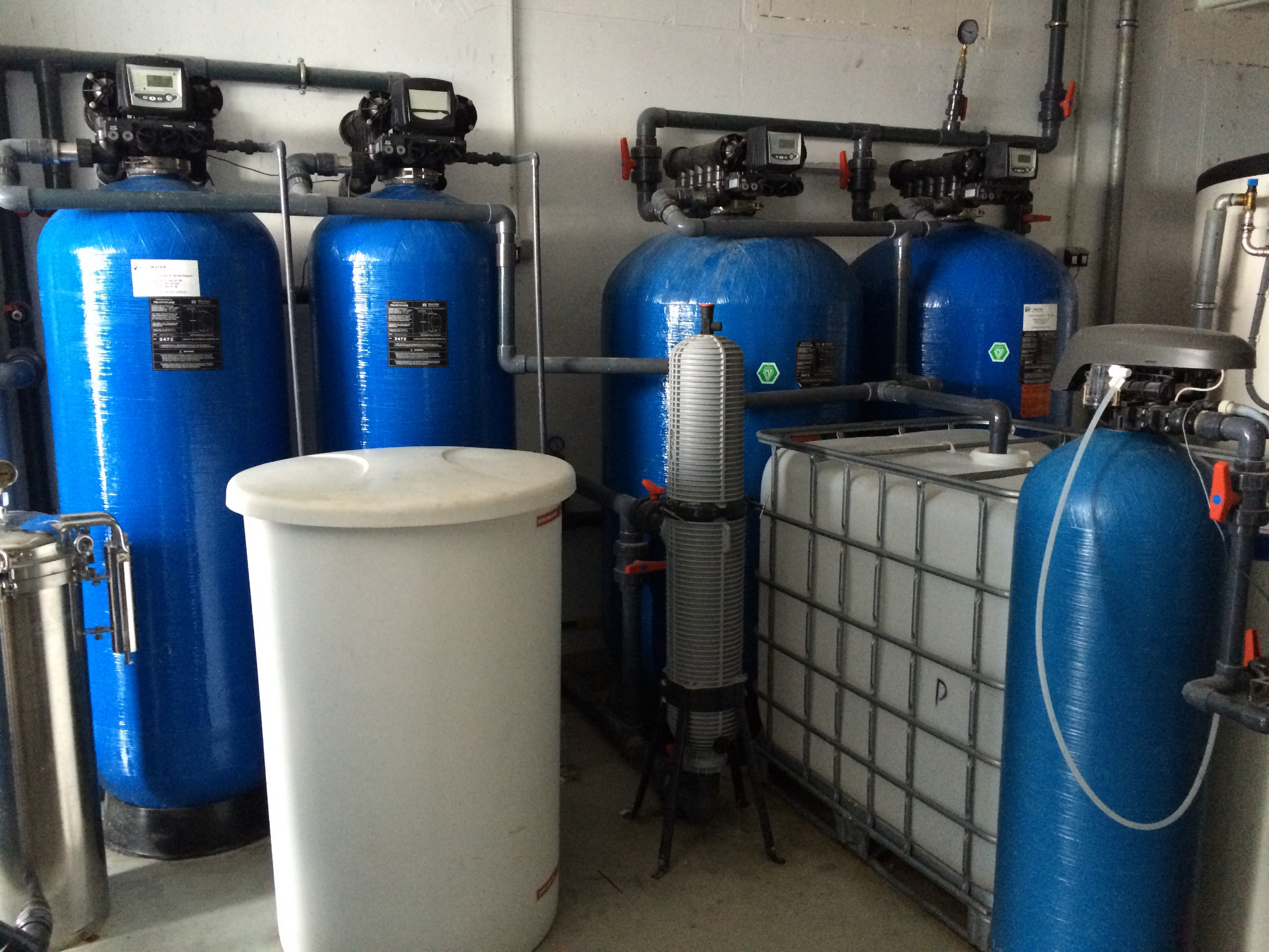 Zestaw urządzeń do filtracji wody w pomieszczeniu technicznym: niebieskie zbiorniki ciśnieniowe z czarnymi głowicami sterującymi, biały pojemnik z tworzywa, filtr kolumnowy na trójnogu...