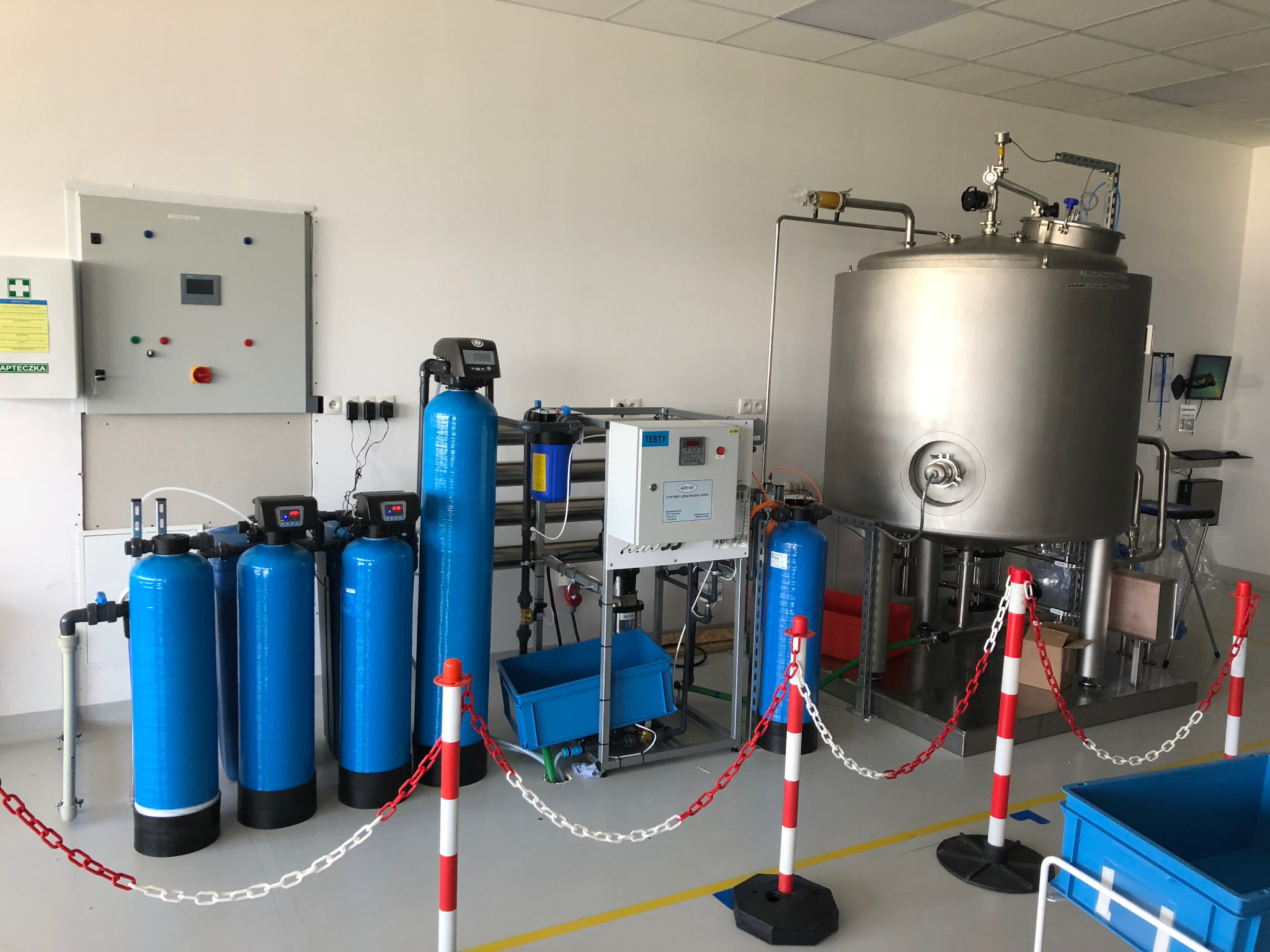 Kompleksowa instalacja przemysłowa z niebieskimi kolumnami filtracyjnymi, zbiornikiem ze stali nierdzewnej i panelem sterowania, zabezpieczona łańcuchem zaporowym w hali produkcyjnej.