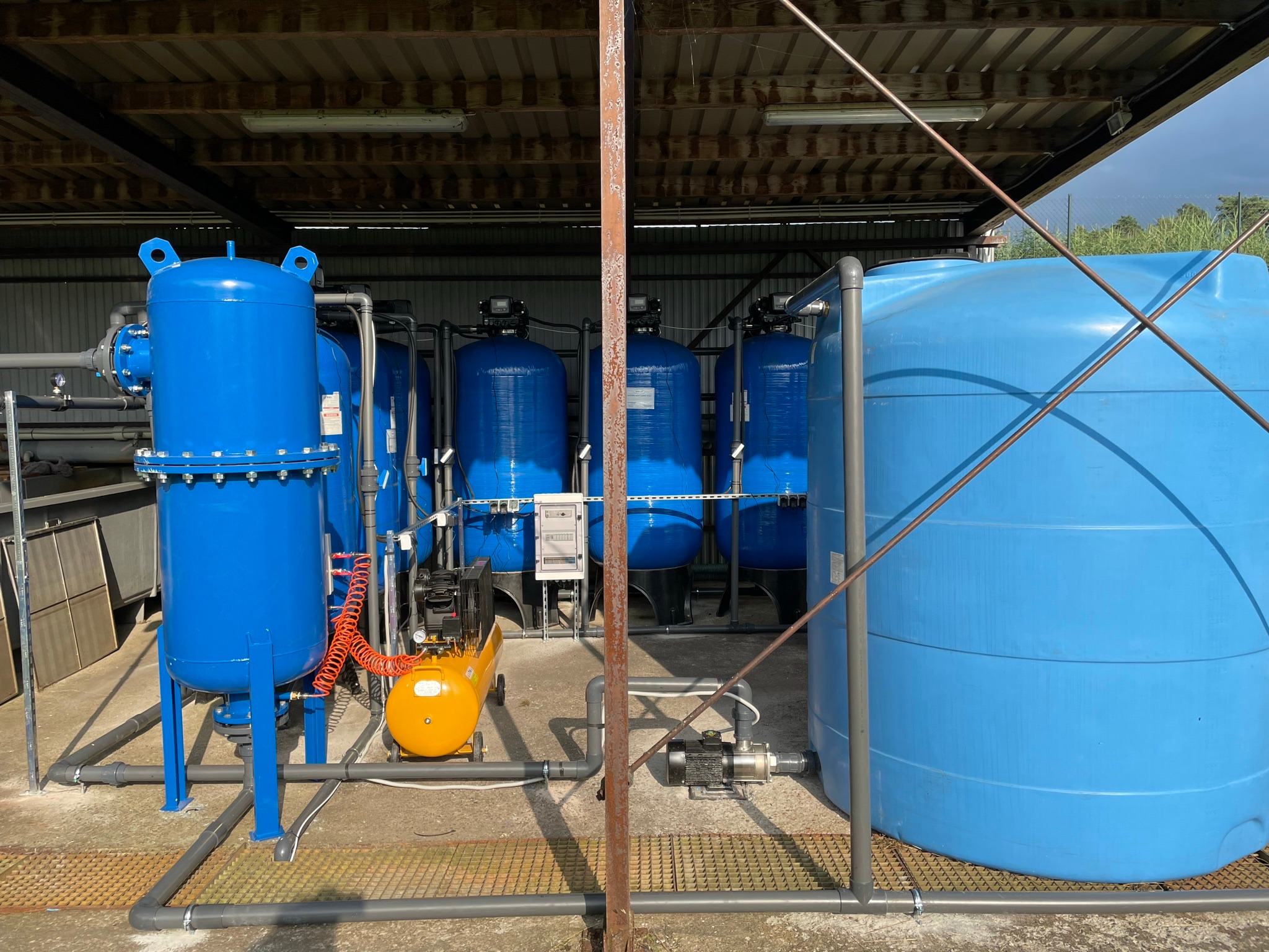 Instalacja uzdatniania wody: niebieskie zbiorniki ciśnieniowe i duży zbiornik na wodę, żółty kompresor i pompa, widoczne rury i elementy instalacji w hali z metalową konstrukcją.